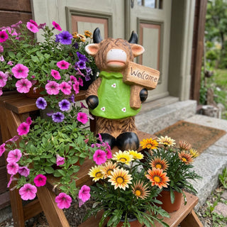 Dekofigur „Galloway-Mädchen Welcome“ (ca. 26 cm hoch) aus Terrakotta, gestaltet als kleine Kuhfigur mit zotteliger Fellstruktur, schwarzen Hörnern und Hufen. Die Figur trägt ein grünes Kleid mit kleinen Blumenmotiven und hält ein Holzschild mit der Aufschrift „Welcome“ in der Hand. Sie steht auf einem dekorativen Holzregal vor einem Hauseingang, umgeben von bunten Blumentöpfen mit pinken, lilafarbenen und gelb-orangen Blüten. Im Hintergrund sind eine Haustür, eine Steintreppe und  Gartenpflanzen zu sehen.