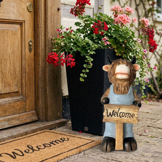 Dekofigur „Galloway-Junge Welcome“ (ca. 41 cm hoch) aus Terrakotta, gestaltet als größere Kuhfigur mit zotteliger Fellstruktur, schwarzen Hörnern und Hufen. Die Figur trägt eine hellblaue Latzhose und hält ein Holzschild mit der Aufschrift „Welcome“ in den Händen. Sie steht auf einem gepflasterten Eingangsbereich neben einer Fußmatte mit der Aufschrift „Welcome“. Im Hintergrund befinden sich eine Holztür sowie ein hoher schwarzer Pflanzkübel mit hängenden grünen Pflanzen und roten sowie rosafarbenen Blüten.