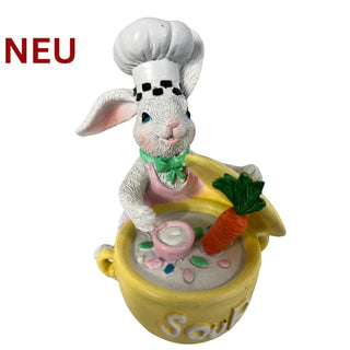 Farbige Dekofigur eines Osterhasen im Kochoutfit mit weißer Kochmütze und grüner Schleife. Der Hase rührt in einem gelben Topf mit Suppe, aus dem eine orangefarbene Karotte ragt. Kleine Details wie Löffel und Gemüsestücke sind sichtbar. Höhe der Figur etwa 12 cm, freigestellt auf hellem Hintergrund.