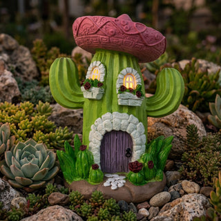 Gartenfigur „Wichtelhaus Kaktus“ (ca. 21 cm) mit Solar-LED, grünes Kaktushaus mit rosa Dach und leuchtenden Fenstern, dekoriert mit kleinen Blüten, steht zwischen Sukkulenten und Steinen.