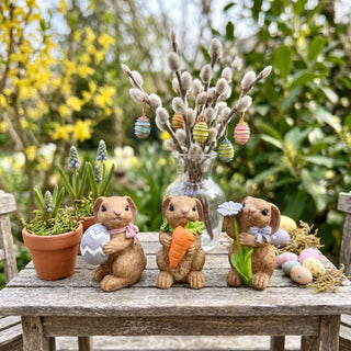 Osterhasen-Set (3-teilig, je ca. 8 cm), drei kleine Hasenfiguren jeweils mit Ei, Möhre und Blume in den Händen. Arrangiert auf einem Holztisch im Garten, umgeben von Blumentöpfen, dekorierten Ostereiern und Frühlingsblüten.