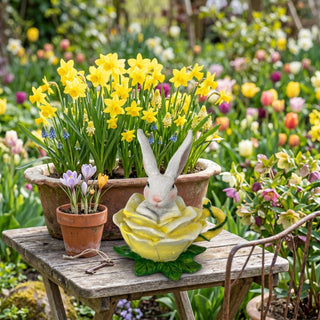 Dekofigur „Osterhase mit Blüte“ (ca. 12 cm), kleiner Hase sitzend in einer gelben Blüte. Arrangiert auf einem Holztisch im Garten vor einem Blumentopf mit gelben Narzissen und bunten Frühlingsblumen.