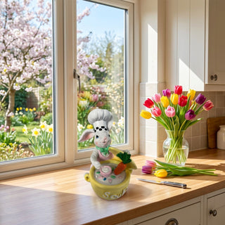 Dekofigur „Osterhase Koch“ (ca. 12 cm), kleiner Hase mit Kochmütze, rührt in einer gelben Schüssel mit Möhre. Steht auf einer Küchenarbeitsplatte neben Tulpen und vor einem Fenster mit Blick in einen Frühlingsgarten.