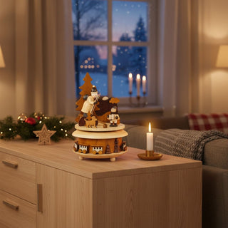 Spieluhr „Bescherung“ (ca. 19 cm) aus naturbelassenem Holz, mit detailreicher Weihnachtsszene aus Figuren um einen Tannenbaum. Die drehbare Plattform zeigt Kinder, Tiere und Geschenke; im Sockel sind kleine Häuser und Zierelemente eingearbeitet. Die Spieluhr steht auf einem Holzmöbel in einer gemütlichen Wohnstube; im Hintergrund ein Fenster mit verschneiter Winterlandschaft sowie Kerzen, die eine warme, festliche Atmosphäre schaffen.