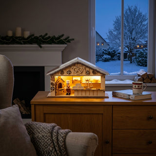 LED-Weihnachtsmarkthütte „Weihnachtsbäckerei“ (27 cm) mit Timer, naturfarbene Holzszene mit Bäckerfigur und Backofen, warm beleuchtet auf einer Kommode vor winterlicher Fensterkulisse platziert.