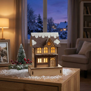 LED-Stadthaus aus Holz (14 cm) mit schneebedecktem Dach und warm leuchtenden Fenstern, dekorativ auf einer Kommode vor winterlicher Fensterkulisse platziert.