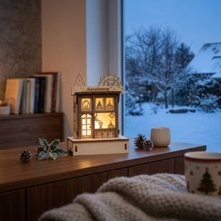 LED-Weihnachtshaus „Spielzeugladen“ (16 cm) aus Holz mit warm beleuchteten Fenstern und detailreicher Szenerie, dekorativ auf einer Kommode vor winterlicher Fensterkulisse platziert.