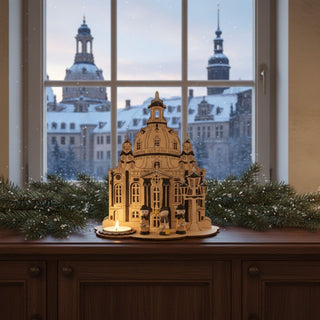 Kerzenhalter aus naturfarbenem Holz für ein Teelicht mit Motiv „Dresdener Frauenkirche“, dekorativ auf einer Fensterbank platziert. Das detailreiche Design zeigt die Frauenkirche mit filigranen Elementen und kleinen Figuren im Vordergrund. Links ist ein Teelicht eingesetzt, dessen warmes Licht die Szene beleuchtet. Im Hintergrund ist durch das Fenster die verschneite Dresdner Altstadt mit Kuppeln bei Abenddämmerung zu sehen; Tannenzweige ergänzen die festliche Atmosphäre.