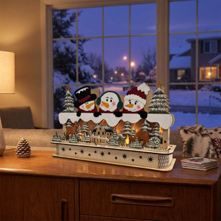 LED-Dekoration „Schneefreunde“ (52 cm) mit Timer, farbige Holzszene mit drei Schneemännern und winterlichem Dorfmotiv, warm beleuchtet auf einer Kommode vor verschneiter Fensterkulisse platziert.