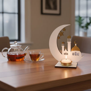 Kerzenhalter für ein Teelicht in Weiß-Gold mit Motiv „Zauber des Orient“, dekoriert auf einem Holztisch. Das Design zeigt eine stilisierte Moschee mit goldener Kuppel und Türmen, eingerahmt von einem großen Halbmond mit Sternen. Ein Teelicht in einer goldenen Halterung sorgt für warmes Licht und hebt die feinen Details hervor. Daneben stehen eine Glaskanne und eine Tasse mit Tee; im Hintergrund ein ruhiger, modern eingerichteter Wohnraum mit Fenstern und Pflanzen.