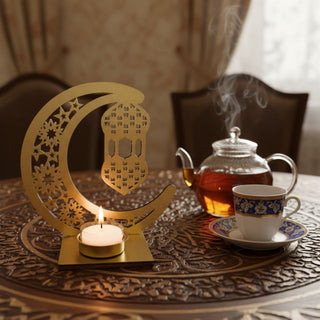 Kerzenhalter für ein Teelicht in goldener Ausführung mit Motiv „Zauber des Orient“, dekoriert auf einem verzierten Tisch. Das kunstvolle Design zeigt einen großen Halbmond mit floralen Mustern und eine darin hängende Laterne. Ein Teelicht in goldener Fassung sorgt für warmes Licht und hebt die filigranen Details hervor. Daneben stehen eine Glaskanne mit dampfendem Tee und eine Tasse mit dekorativem Muster; im Hintergrund ein gemütlicher Wohnraum mit Stühlen.
