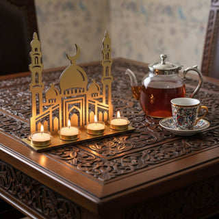 Kerzenhalter für vier Teelichte in goldener Ausführung mit Motiv „Zauber des Orient“, dekoriert auf einem kunstvoll verzierten Holztisch. Das Design zeigt eine stilisierte orientalische Architektur mit Kuppel, Minaretten und Rundbögen. Vier Teelichter in goldenen Haltern sorgen für warmes Licht und betonen die filigranen Details. Daneben stehen eine Glaskanne mit Tee und eine verzierte Tasse; im Hintergrund sind Möbel und eine orientalisch anmutende Umgebung zu sehen.