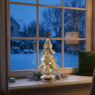 LED-Weihnachtsdeko aus Mercury-Glas „Weihnachtsbaum“ (26 cm), warm leuchtend auf einer Fensterbank platziert. Der dekorative Glasbaum mit strukturierter Oberfläche und Stern an der Spitze strahlt ein goldenes Licht aus. Im Hintergrund ein Fenster mit verschneiter Winterlandschaft, ergänzt durch weihnachtliche Details wie Tannenzweig, Schleife und Teelicht sowie ein gemütlicher Innenraum mit Kissen und Lampe.