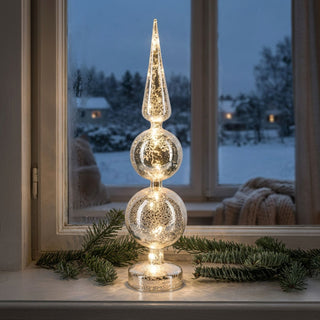 LED-Weihnachtsdeko aus Mercury-Glas „Spitze“ (43 cm), elegant und warm leuchtend auf einer Fensterbank platziert. Die schlanke, mehrteilige Glasform mit Kugel-Elementen und spitz zulaufender Krone zeigt eine strukturierte Oberfläche im schimmernden Metallic-Look. Umgeben von Tannenzweigen, vor einem Fenster mit verschneiter Winterlandschaft und dezent beleuchteten Häusern im Hintergrund, sorgt die Dekoration für eine festliche und gemütliche Atmosphäre.