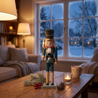 Nussknacker, 25 cm groß, aus blau lasiertem Holz, mit schwarzem Zylinder, weißem Bart und Schnurrbart, in klassischer Uniform mit goldenen Zierelementen und Stab, dekorativ auf einem Tisch vor winterlicher Fensterkulisse mit Kerzenlicht, Tannenzweig und warm beleuchtetem Wohnambiente platziert.