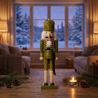 Nussknacker, 25 cm groß, aus grün lasiertem Holz, mit grünem Hut, weißem Bart und Schnurrbart, in verzierter Uniform, dekorativ auf einem Tisch mit Tannenzweigen, Zapfen und Kerzenlicht vor verschneiter Fensterkulisse im warmen Wohnambiente platziert.