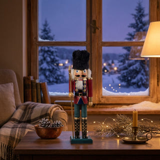 Nussknacker, 25 cm groß, aus rot lasiertem Holz, mit schwarzer Fellmütze, weißem Bart und Schnurrbart, in klassischer Uniform mit Zierelementen und Stab in der Hand, dekorativ auf einem Tisch vor winterlicher Fensterkulisse mit Kerzenlicht und warmem Wohnambiente platziert.