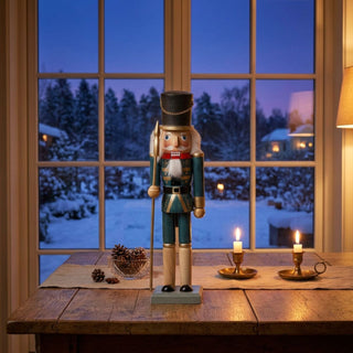 Nussknacker blau lasiert (39 cm) mit Hut, Bart und Lanze, steht auf einem Holztisch. Umgeben von Kerzenhaltern, Zapfen und winterlicher Dekoration, vor einem großen Fenster mit verschneiter Landschaft in gemütlichem Wohnambiente.