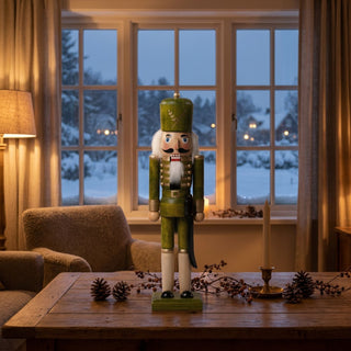 Nussknacker grün lasiert (39 cm) mit Hut, Bart und Säbel, steht auf einem Holztisch. Umgeben von Kerzenhalter, Zapfen und Zweigen, vor einem gemütlichen Wohnzimmer mit Sessel und verschneiter Winterlandschaft am Fenster.