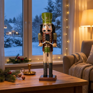 Nussknacker „Trommler“ grün lasiert (39 cm) mit Trommel und Trommelstöcken, steht auf einem Holztisch. Umgeben von Kerzenhalter, Tannenzweigen und Nüssen, vor einem gemütlichen Wohnzimmer mit Lichterkette und verschneiter Winterlandschaft am Fenster.