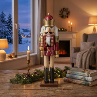 Nussknacker „König“ rot lasiert (39 cm) mit goldener Krone, Zepter und weißem Bart, steht auf einem Holztisch. Umgeben von Kerze, Tannenzweigen, Zapfen und Büchern, vor einem gemütlichen Wohnzimmer mit Kamin und verschneiter Winterlandschaft am Fenster.