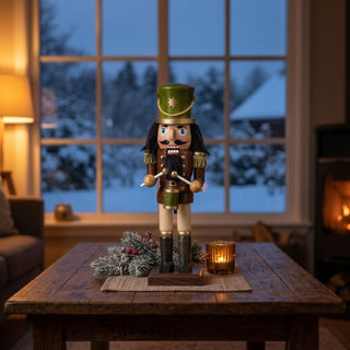 Nussknacker „Trommler“, 25 cm groß, aus braun lasiertem Holz, mit grünem Zylinder, schwarzem Bart und Schnurrbart, in einer Trommleruniform mit goldenen Details sowie Trommelstöcken in den Händen, dekorativ auf einem Tisch vor winterlich beleuchtetem Fenster und Kerzenlicht platziert.