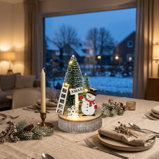 LED-Weihnachtsdekoration „Schneemann“, farbig (12 cm) auf Holzsockel, mit beleuchtetem Tannenbaum, „XMAS“-Schild und Leiter, dekorativ auf einem gedeckten Tisch vor winterlicher Fensterkulisse platziert.