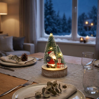 LED-Weihnachtsdekoration „Weihnachtsmann“ (12 cm) mit warmem Licht auf einem Holzsockel, dekorativ auf einem gedeckten Tisch platziert. Die Szene zeigt eine Weihnachtsmannfigur mit Geschenken vor kleinen, beleuchteten Tannenbäumen. Im Vordergrund befinden sich Teller, Besteck, Glas und Servietten mit winterlicher Dekoration, während im Hintergrund ein gemütlicher Wohnraum mit Sofa und verschneiter Landschaft vor dem Fenster zu sehen ist.