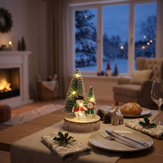 LED-Weihnachtsdekoration „Schneemann mit Kind“ (12 cm) mit warmem Licht auf einem Holzsockel, dekorativ auf einem gedeckten Tisch platziert. Die Szene zeigt zwei Schneemannfiguren vor kleinen, beleuchteten Tannenbäumen und sorgt für eine festliche Atmosphäre. Im Vordergrund befinden sich Geschirr, Besteck und weihnachtlich dekorierte Servietten, während im Hintergrund ein gemütliches Wohnzimmer mit Kaminfeuer, Sessel und verschneiter Winterlandschaft vor dem Fenster zu sehen ist.