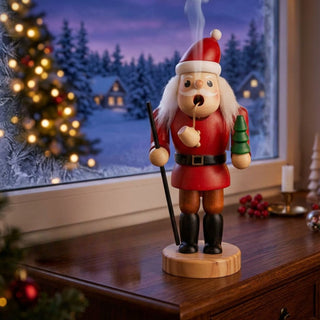 Farbiges Räuchermännchen „Weihnachtsmann“ (13 cm) aus Holz mit roter Kleidung, Weihnachtsmütze, Pfeife und kleinem Tannenbaum in der Hand. Aus der Pfeife steigt feiner Rauch auf. Die Figur steht auf einem Holzmöbel vor einem Fenster mit verschneiter Winterlandschaft und beleuchteten Häusern. Im Hintergrund sorgen ein geschmückter Weihnachtsbaum, Kerzen und warme Lichter für eine festliche und gemütliche Weihnachtsatmosphäre.