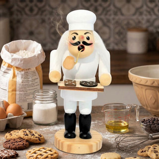 Farbiges Räuchermännchen „Bäcker“ (13 cm) aus Holz mit weißer Bäckerkleidung, hoher Kochmütze und Pfeife. Auf einem Tablett präsentiert die Figur verschiedene Plätzchen. Aus der Pfeife steigt feiner Rauch auf. Das Räuchermännchen steht auf einer Arbeitsfläche in einer Backstube, umgeben von Backzutaten wie Mehl, Eiern, Zucker, Öl, Schokoladenstückchen und einer Rührschüssel. Frisch gebackene Kekse und die warme Küchenszene unterstreichen die gemütliche Atmosphäre.