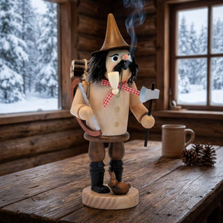 Naturfarbenes Räuchermännchen „Holzfäller“ (32 cm) aus Holz mit spitzem Hut, Pfeife, Axt und Handsäge. Aus der Pfeife steigt feiner Rauch auf. Auf dem Rücken trägt die Figur ein Bündel Holzscheite, zu ihren Füßen sitzt ein kleiner Hase. Das Räuchermännchen steht auf einem rustikalen Holztisch in einer gemütlichen Blockhütte. Durch die Fenster ist eine verschneite Waldlandschaft zu sehen. Die warme Holzoptik und die winterliche Kulisse unterstreichen den traditionellen Charakter und die behagliche Atmosphäre