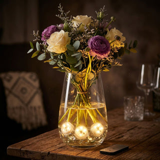 LED-Teelichter (6er-Set) wasserdicht mit Fernbedienung, leuchten warmweiß in einer Glasvase zwischen Blumenstielen, dekorativ auf einem Holztisch platziert neben Fernbedienung und Blumenstrauß.