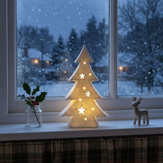 LED Weihnachtsbaum aus Porzellan (21 cm) in Weiß, warmweiß beleuchtet, mit ausgestanzten Sternen und Lichtpunkten, als stilvolle Fensterbankdekoration vor winterlicher Schneelandschaft.
