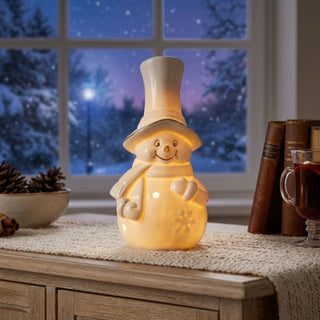 LED Schneemann mit Zylinder (16 cm) aus Porzellan, warmweiß beleuchtet, mit ausgestanzten Lichtpunkten und Schneeflockenmotiv, als stimmungsvolle Tischdekoration vor winterlicher Fensterkulisse.