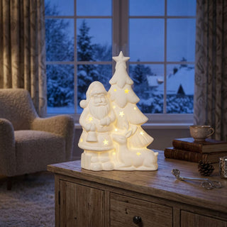LED-Weihnachtsbaum mit Weihnachtsmann (18 cm) aus Porzellan, mit warm leuchtenden Sternausschnitten, dekorativ auf einer Kommode vor winterlicher Fensterlandschaft platziert.