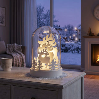 LED Glasdome „Hirsche“ (18 cm) mit klarer Glaskuppel, warmweiß beleuchtet, filigrane Silhouetten von Hirschen und Tannenbäumen im Inneren, dekorativ auf Sockel platziert als stimmungsvolle Winterdekoration im gemütlichen Wohnambiente mit verschneiter Landschaft im Hintergrund.