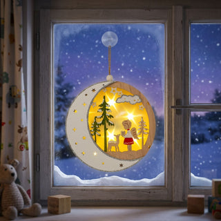 Fenster mit verschneiter Winterlandschaft bei Nacht. In der Mitte hängt ein runder, warm beleuchteter Holz-Fensteranhänger mit Halbmondrahmen und kleinen Sternen. Im Inneren sind eine stilisierte Szene mit Tannenbäumen, einem Engel und einem Reh vor leuchtendem Sternenhintergrund zu sehen. Der Anhänger ist mit Schnur und Saugnapf befestigt; auf der Fensterbank liegen Holzklötze, links steht ein Stofftier.