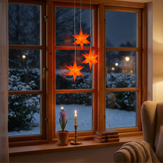 Drei rote LED-Adventssterne mit je 12 cm Durchmesser hängen an weißen Kabeln vor einem Holzfenster. Die leuchtenden Sterne sorgen für eine warme Weihnachtsstimmung, während draußen eine verschneite Winterlandschaft zu sehen ist. Auf der Fensterbank stehen eine Kerze, eine Topfpflanze und einige Bücher. Das 3er-Set von HGD eignet sich als stimmungsvolle Fensterdekoration in der Advents- und Weihnachtszeit.