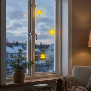 Drei gelb leuchtende LED-Adventssterne mit einem Durchmesser von jeweils 12 cm hängen an feinen Kabeln vor einem Fenster. Die Sterne strahlen warmes Licht aus und setzen winterliche Akzente in der Wohnraumdekoration. Auf der Fensterbank steht ein Topf mit Tannenzweigen, daneben liegen Bücher. Im Hintergrund ist eine verschneite Landschaft zu sehen.
