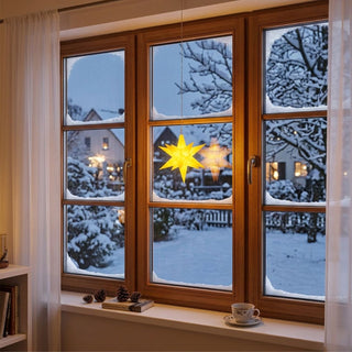LED Adventsstern von HGD in Gelb mit einem Durchmesser von 12 cm. Der leuchtende Weihnachtsstern hängt an einer feinen Schnur vor einem großen Holzfenster und strahlt ein warmes, gelbes Licht aus. Durch das Fenster ist eine verschneite Winterlandschaft mit schneebedeckten Bäumen, Häusern und Gartenflächen zu sehen. Auf der Fensterbank stehen dekorative Tannenzapfen, ein Buch und eine Tasse, die eine gemütliche Wohnatmosphäre schaffen. 