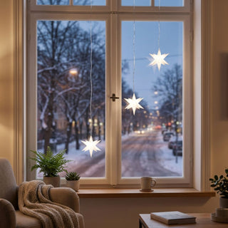Drei weiß leuchtende LED-Adventssterne mit einem Durchmesser von jeweils 12 cm hängen an dünnen Kabeln vor einem großen Fenster. Die Sterne sorgen für eine festliche Atmosphäre und setzen helle Lichtakzente im Wohnraum. Auf der Fensterbank stehen Zimmerpflanzen und eine Tasse, daneben befindet sich ein gemütlicher Sessel mit Strickdecke. Durch das Fenster ist eine verschneite Straße mit winterlicher Abendstimmung zu sehen.