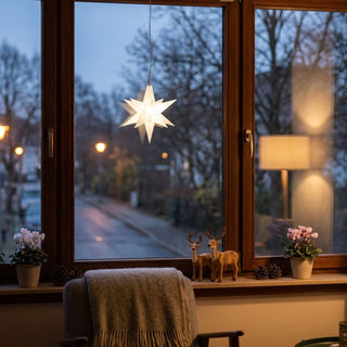 LED Adventsstern von HGD in Weiß mit einem Durchmesser von 12 cm. Der beleuchtete Weihnachtsstern hängt an einer feinen Schnur vor einem großen Holzfenster und strahlt ein warmweißes Licht aus. Im Hintergrund ist eine winterliche Straße mit kahlen Bäumen und Straßenbeleuchtung in der Dämmerung zu sehen. Auf der Fensterbank befinden sich dekorative Figuren von Rentieren, Tannenzapfen und Blumentöpfe, während ein Sessel mit Decke und eine Stehlampe für eine gemütliche Wohnatmosphäre sorgen.