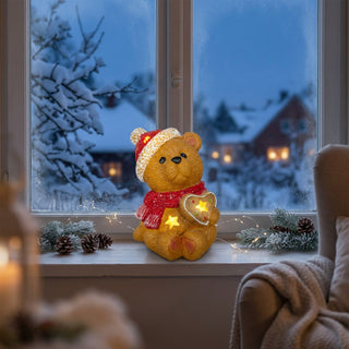 LED-Dekoration „Teddybär mit Herz“ (15 cm) für Batteriebetrieb, warm leuchtend auf einer Fensterbank platziert. Der Keramikbär trägt eine rote Mütze und einen roten Schal und hält ein Herz mit integrierten Sternlichtern. Umgeben von winterlicher Dekoration wie Tannenzweigen, Zapfen und einer feinen Lichterkette, mit Blick auf eine verschneite Landschaft und beleuchtete Häuser im Hintergrund sowie einen gemütlichen Innenraum mit Sessel.