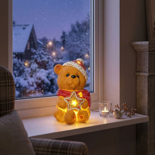 LED-Dekoration „Teddybär mit Stern“ (15 cm) für Batteriebetrieb, warm leuchtend auf einer Fensterbank platziert. Der Keramikbär trägt eine rote Mütze und einen roten Schal und hält einen dekorativen Stern mit integrierten Lichtern. Im Hintergrund ist eine verschneite Winterlandschaft mit Häusern und warmen Lichtern zu sehen, ergänzt durch ein Teelichtglas und kleine Dekorationen auf der Fensterbank sowie ein gemütlicher Innenraum mit Sessel und Ofen.