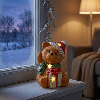 LED-Dekoration „Teddybär mit Geschenk“ (15 cm) für Batteriebetrieb, warm leuchtend auf einer Fensterbank platziert. Der Keramikbär trägt eine rote Mütze und einen grünen Schal und hält ein Geschenk mit roten Band und integrierten Sternlichtern. Im Hintergrund ein winterlicher Blick aus dem Fenster mit verschneiter Landschaft sowie ein gemütlich beleuchteter Innenraum mit Sofa und Stehlampe.