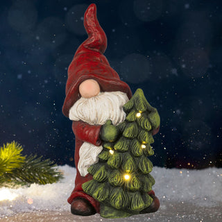 Dekorativer Winterwichtel mit roter, spitzer Mütze und langem weißem Bart, der einen grünen Tannenbaum mit warmweißen Lichtern umarmt. Die Figur steht im Schnee vor einem dunklen, winterlichen Hintergrund mit Schneeflocken und einem Tannenzweig.