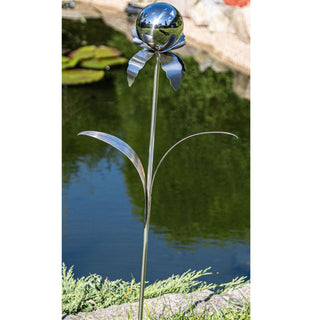 Gartenstecker „Blume“ aus Edelstahl im Außenbereich platziert, mit glänzender Kugel als Blütenkopf und filigranen Metallblättern, steckt am Rand eines Gartenteichs mit Wasserpflanzen und Steinumrandung im Hintergrund.