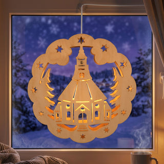 LED‑Fensterbild aus naturfarbenem Holz mit Motiv der Seiffener Kirche, umgeben von Sternen und stilisierten Tannenzweigen. Die warme Beleuchtung hebt die filigranen Ausschnitte hervor; im Hintergrund ist eine verschneite Winterlandschaft vor einem Fenster zu sehen, daneben eine Tasse und Wohnaccessoires.
