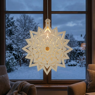 LED‑Fensterbild in Form eines sternförmigen Eissterns aus naturfarbenem Holz, filigran gearbeitet und warm beleuchtet. Mehrschichtiger Stern mit feinen Mustern und kleinen ausgeschnittenen Sternen, aufgehängt vor einem Fenster mit verschneiter Winterlandschaft im Hintergrund; seitlich sind Sessel und Wohntextilien zu sehen.